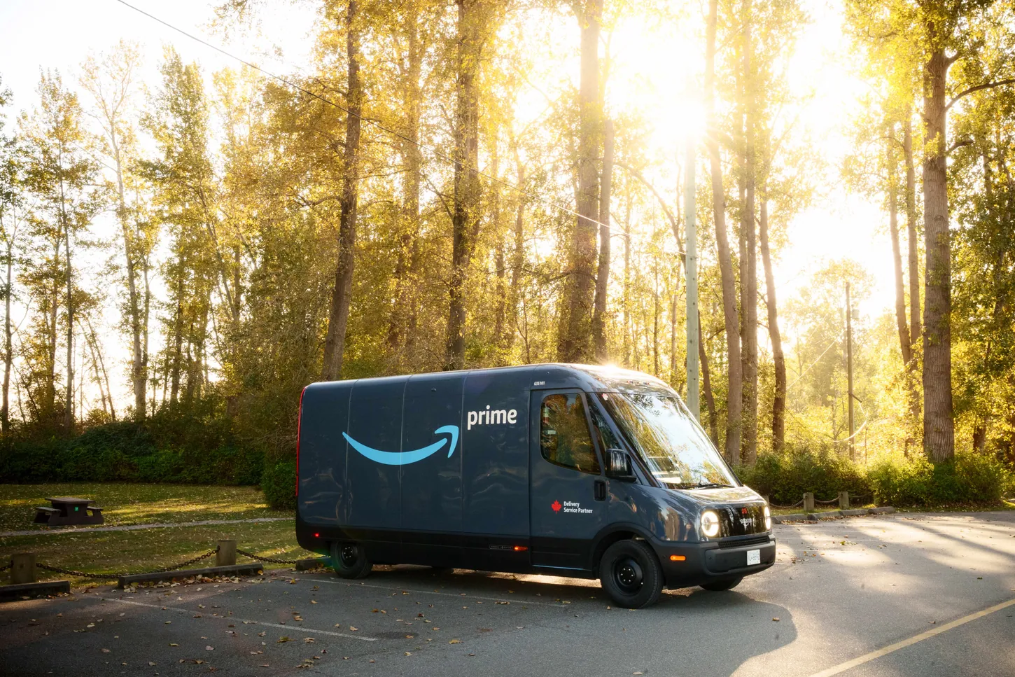 Dark blue Amazon Prime electric delivery van by Rivian parked in a sunlit lot lined with tall trees