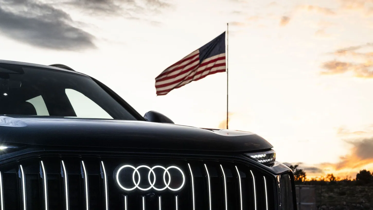 Front of a black Audi Q9 SUV with illuminated four-ring logo and vertical grille lights, an American flag flying against a sunset sky behind it