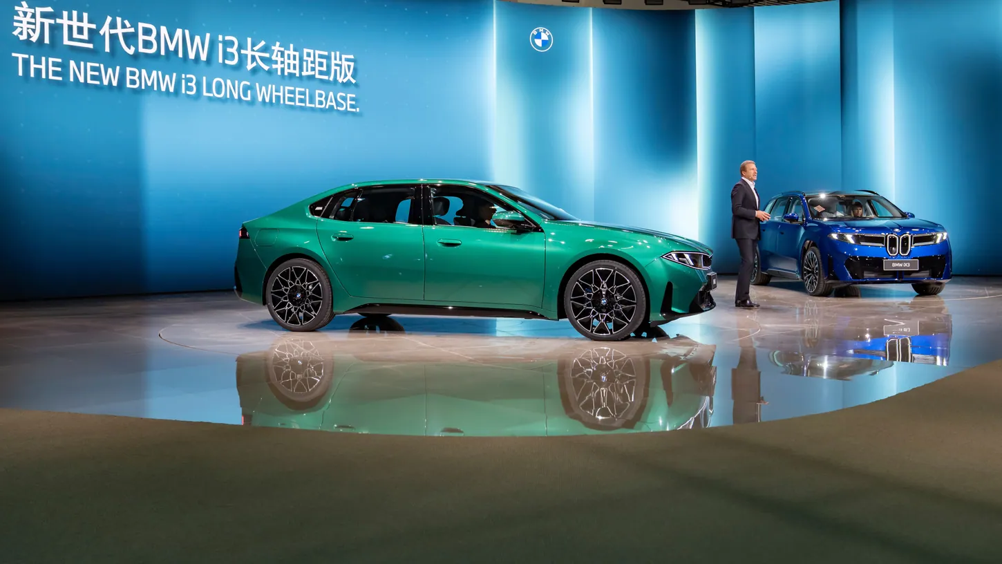 Green BMW i3 Long Wheelbase sedan and blue BMW iX3 SUV on a show stage under a sign reading The New BMW i3 Long Wheelbase