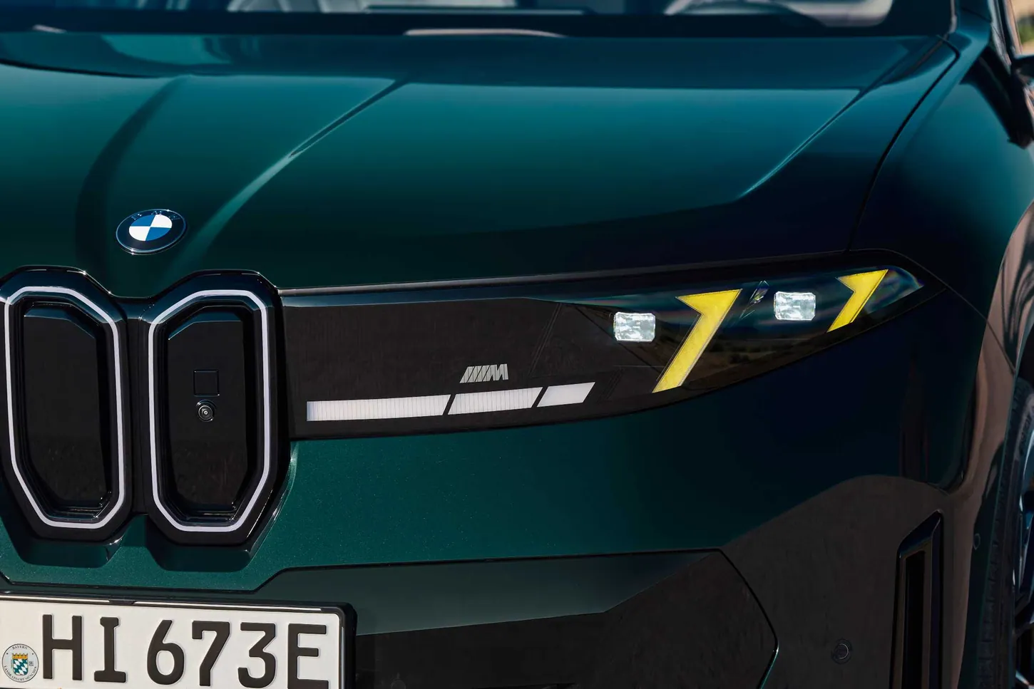 Close-up of the BMW iX3 M60 xDrive's front end showing the yellow headlight, the M badge and the three-element light bar next to the kidney grille