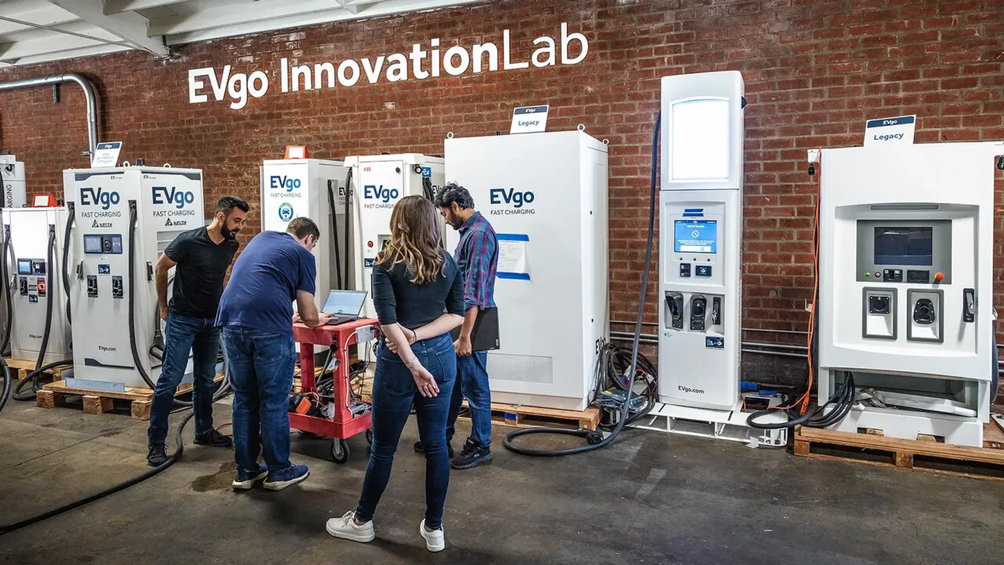 Four engineers at a laptop cart in front of a row of white EVgo fast chargers inside a brick-walled lab marked EVgo Innovation Lab
