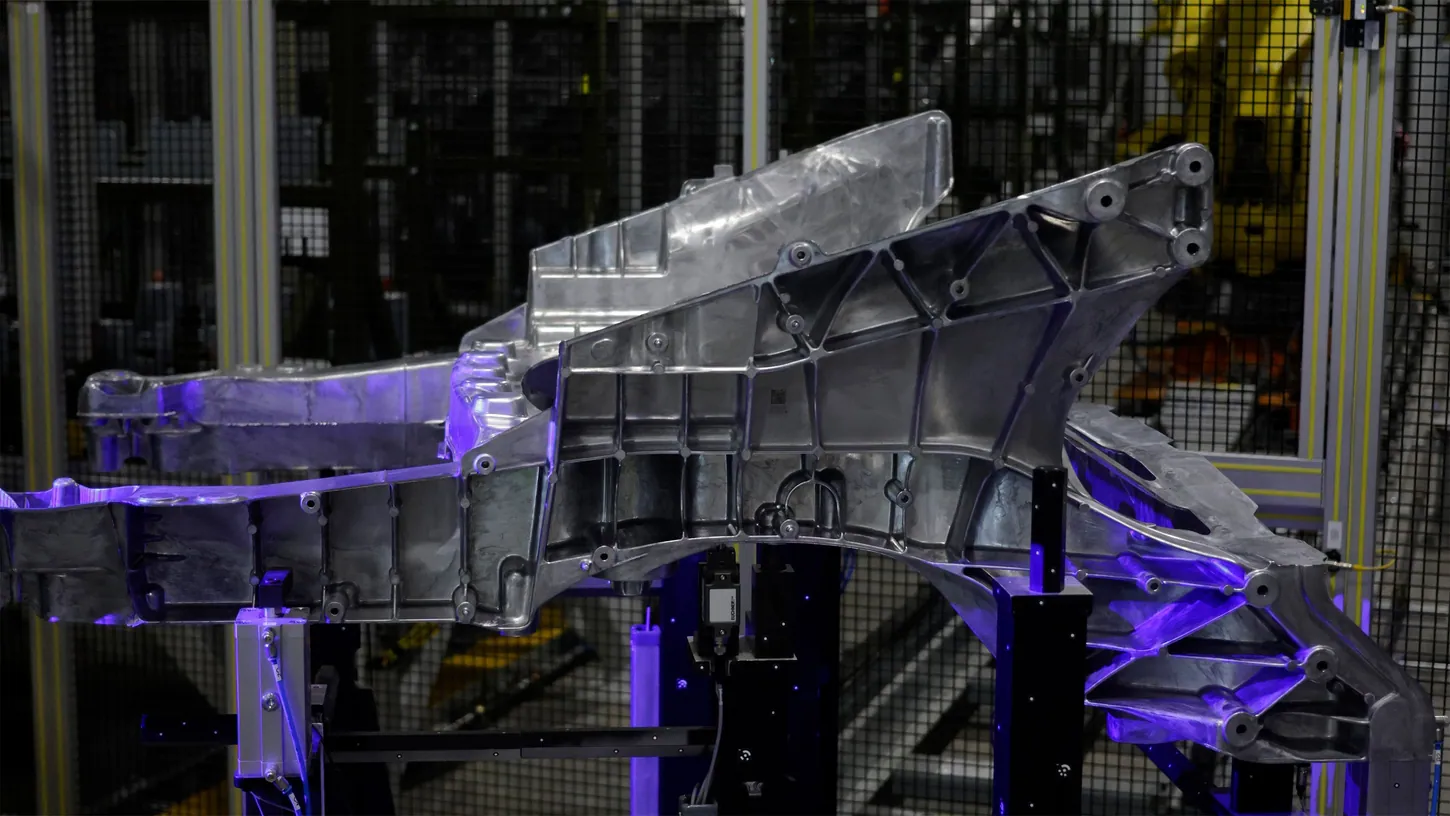 Large raw aluminum casting mounted on black fixtures under purple light inside a caged optical scanning cell