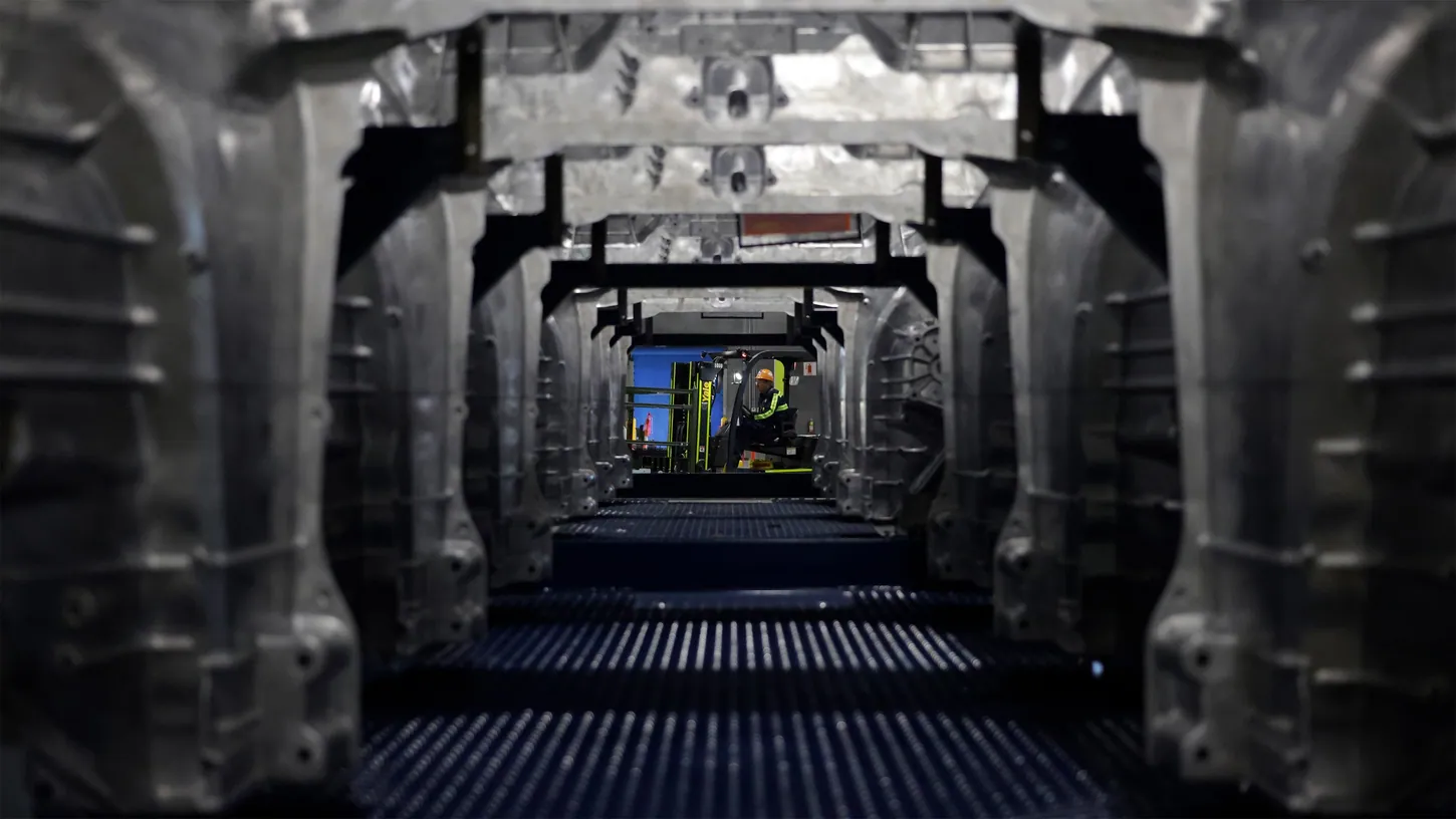 Rows of raw aluminum castings stacked on racks forming a tunnel, with a forklift driver visible at the far end