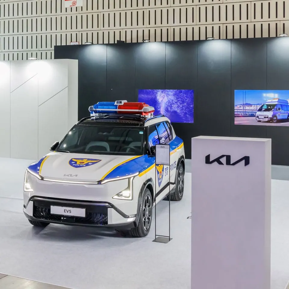 White Kia EV5 SUV in Korean police livery with a roof light bar, parked beside a Kia sign on an exhibition stand