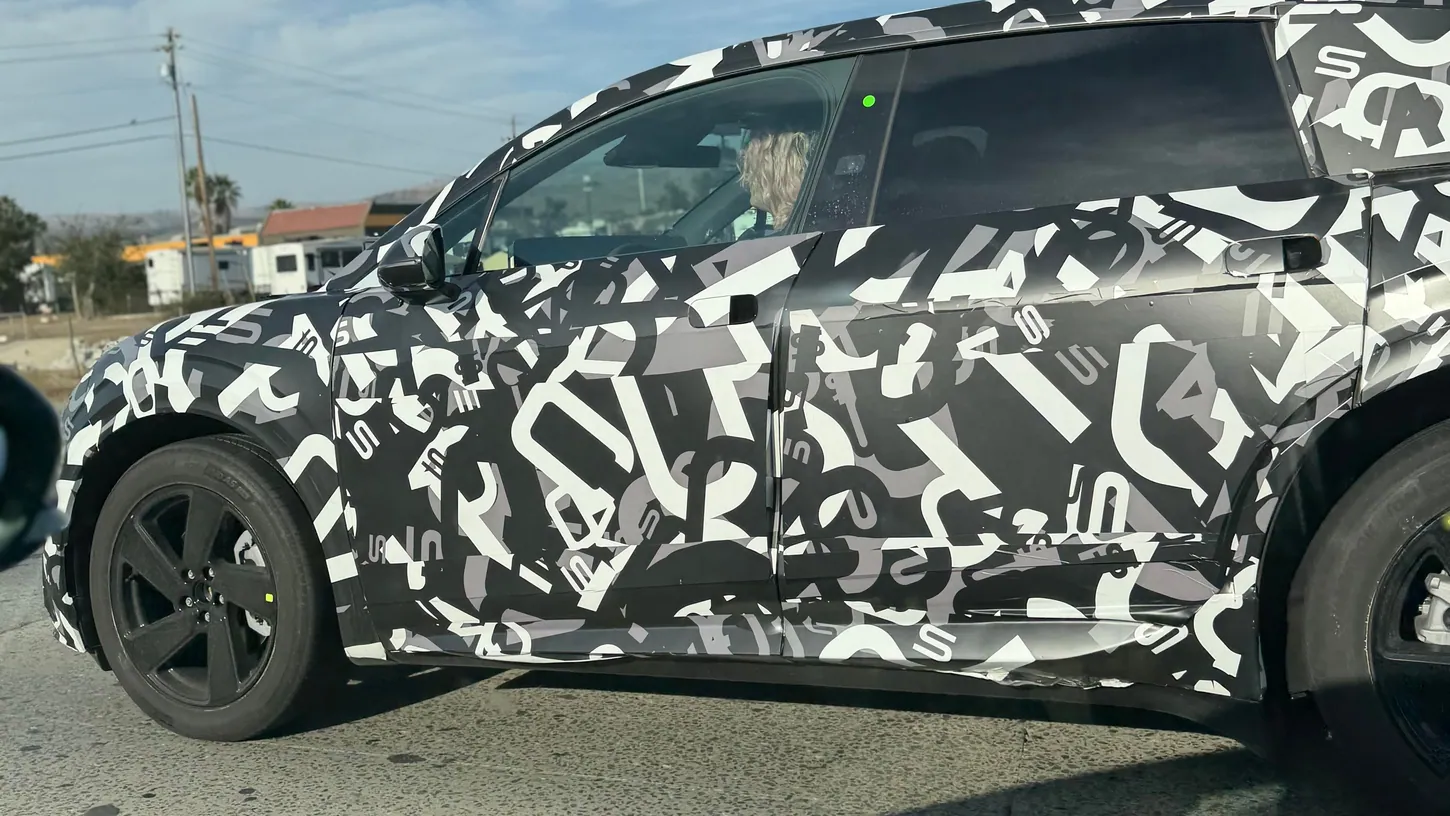Close side view of a camouflaged Lucid Cosmos prototype showing flush door handles, a black five-spoke wheel and the driver behind the window