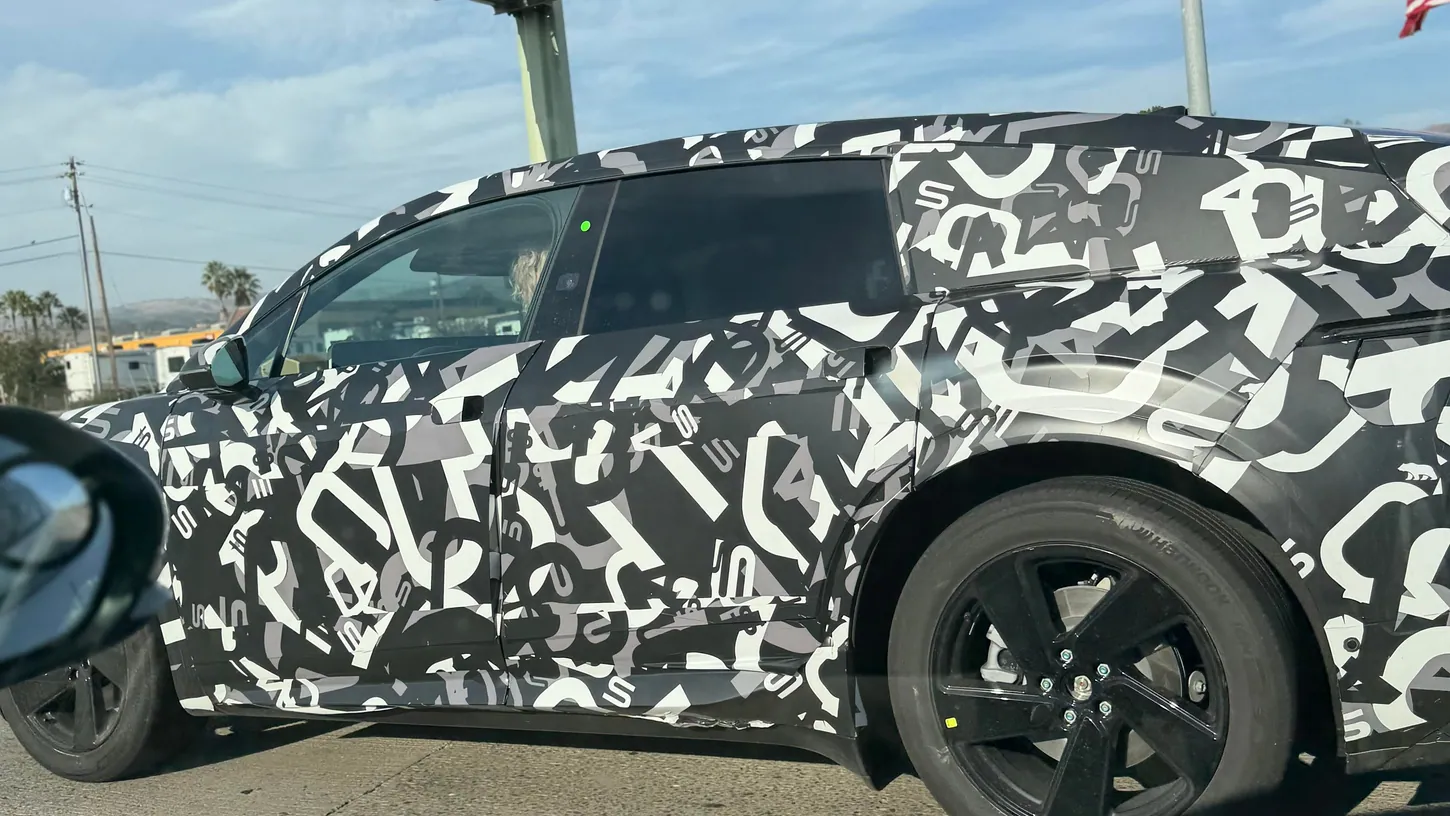Side view of a camouflaged Lucid Cosmos prototype on a highway under an East Dunne Avenue sign, showing black five-spoke wheels and a green light on the B-pillar