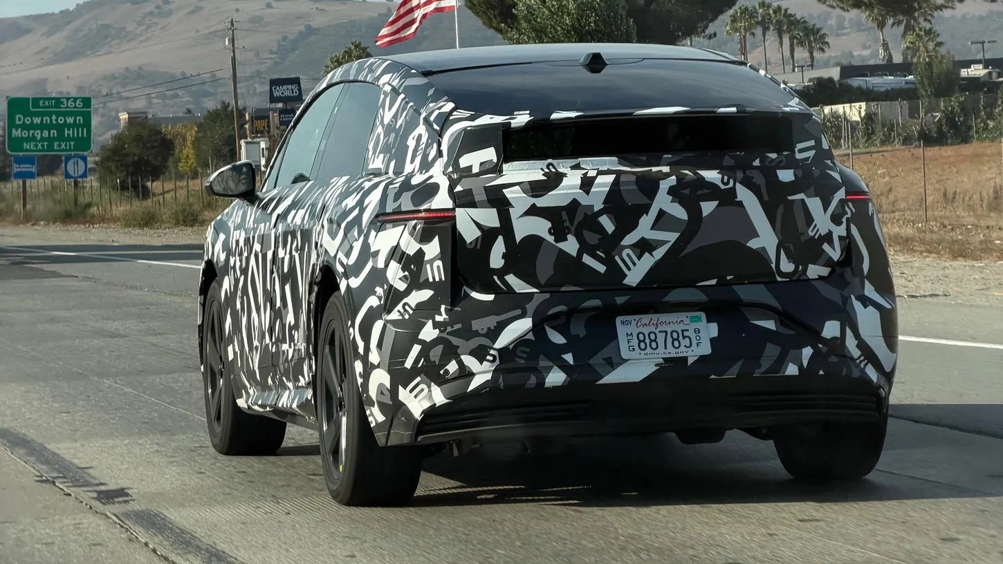 Black and white camouflaged Lucid Cosmos prototype from behind on a highway, with a Downtown Morgan Hill exit sign and an American flag in the background