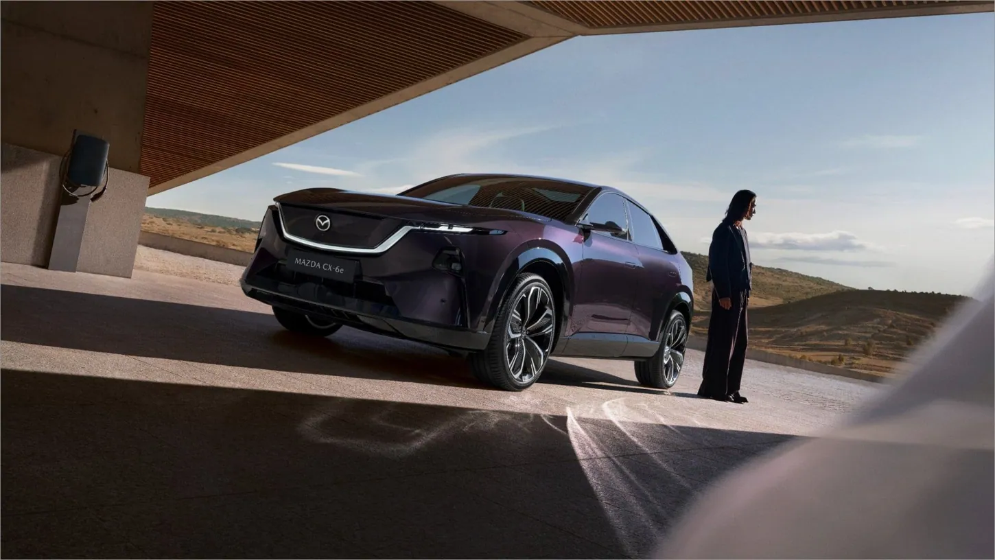 Dark purple Mazda CX-6e parked under a roof next to a wall-mounted charger, with a person standing beside the passenger door