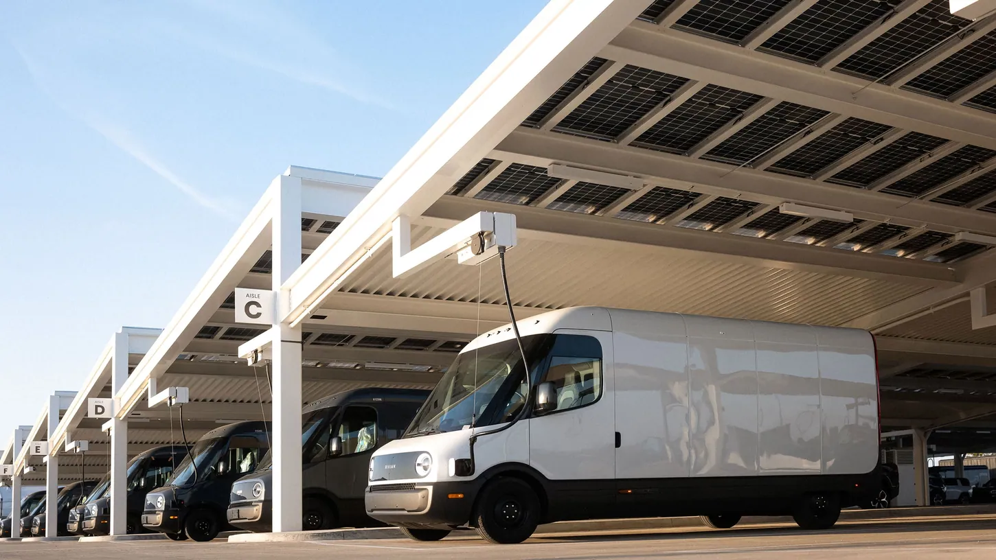 White Rivian Commercial Van plugged in under a solar canopy at a charging depot, with dark Rivian vans lined up behind it