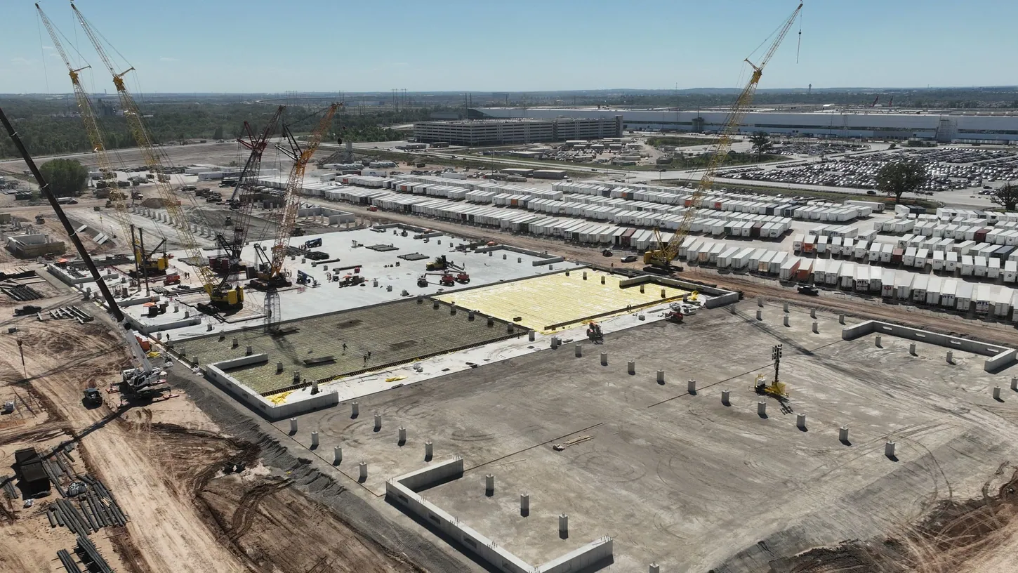 Aerial view of cranes and concrete foundations at a large construction site beside rows of parked trailers and a factory building