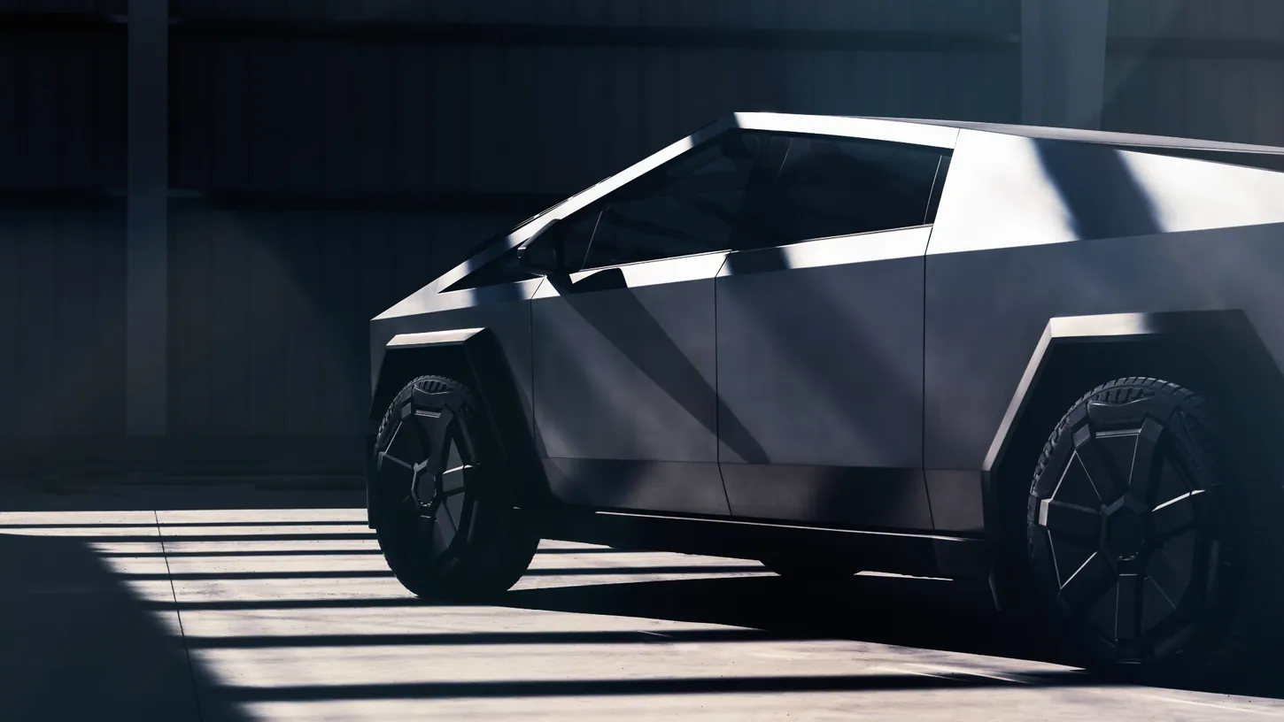 Stainless steel Tesla Cybertruck seen from the front three-quarter angle, parked in a dim warehouse with bars of sunlight falling across its body and the floor