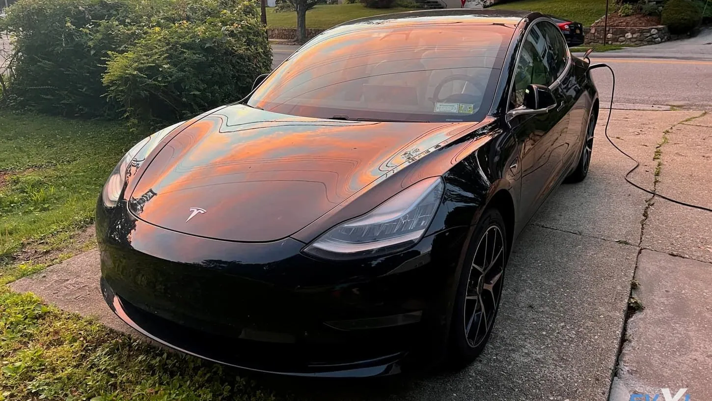 Black Tesla Model 3 parked on a driveway at sunset with a charging cable plugged in