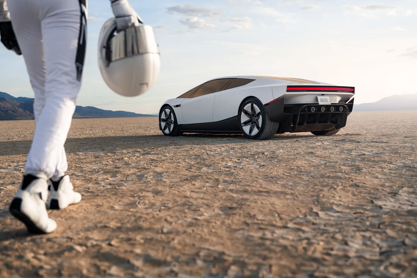 Driver in a white race suit carrying a white helmet walks toward the white Tesla Roadster parked on a dry lakebed