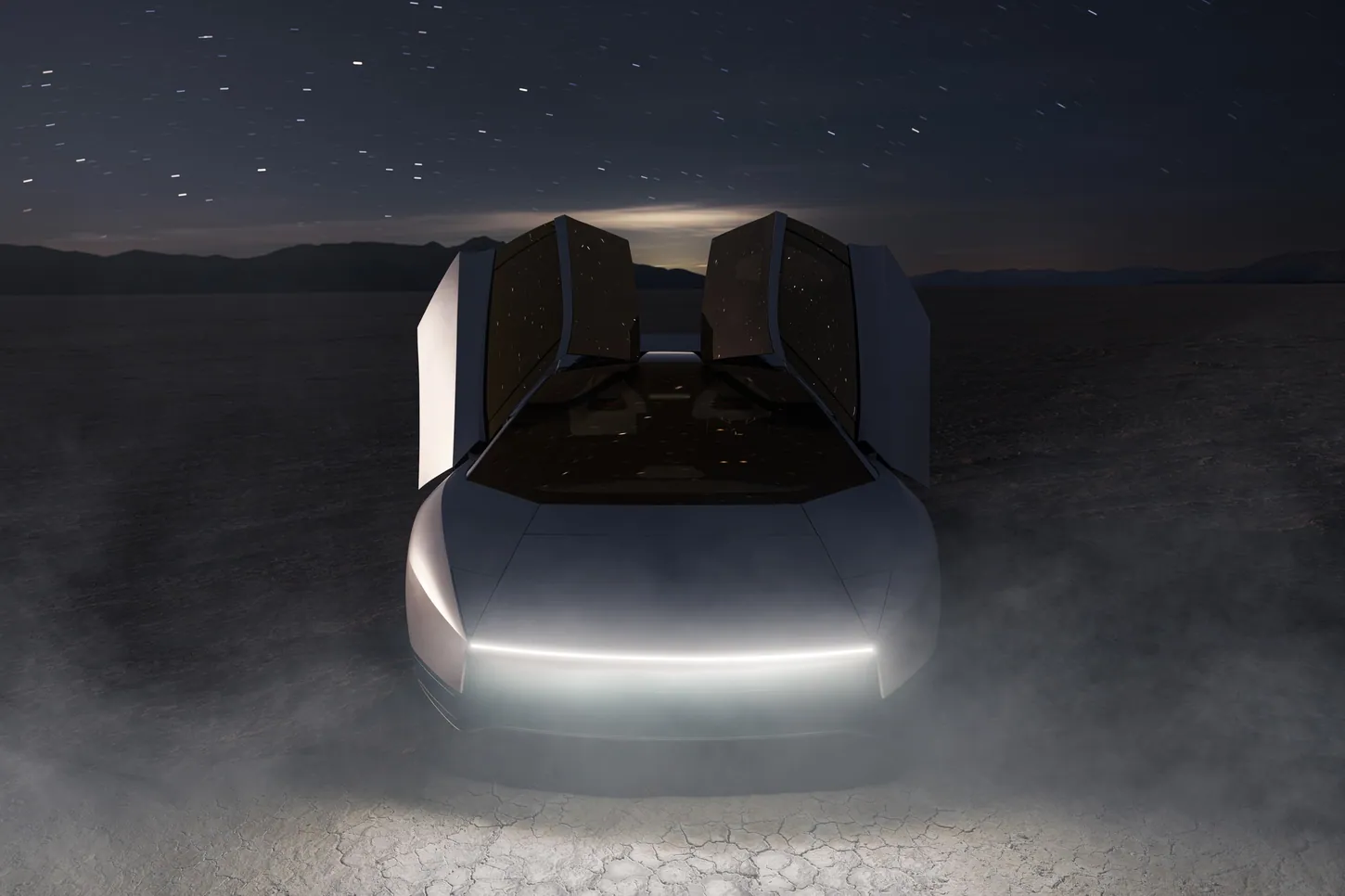 Tesla Roadster seen head-on at night on a dry lakebed, both doors raised and a thin white light bar glowing across the nose