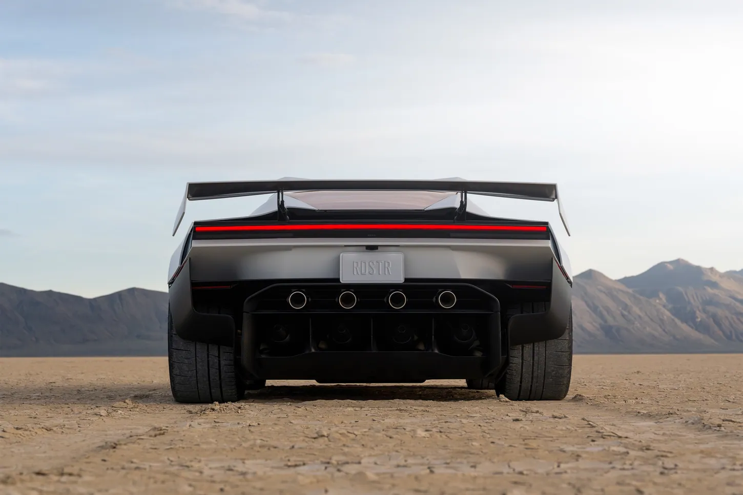 Rear view of the white Tesla Roadster on a dry lakebed, with its wing raised, four thruster nozzles under the RDSTR plate and four fans in the diffuser below