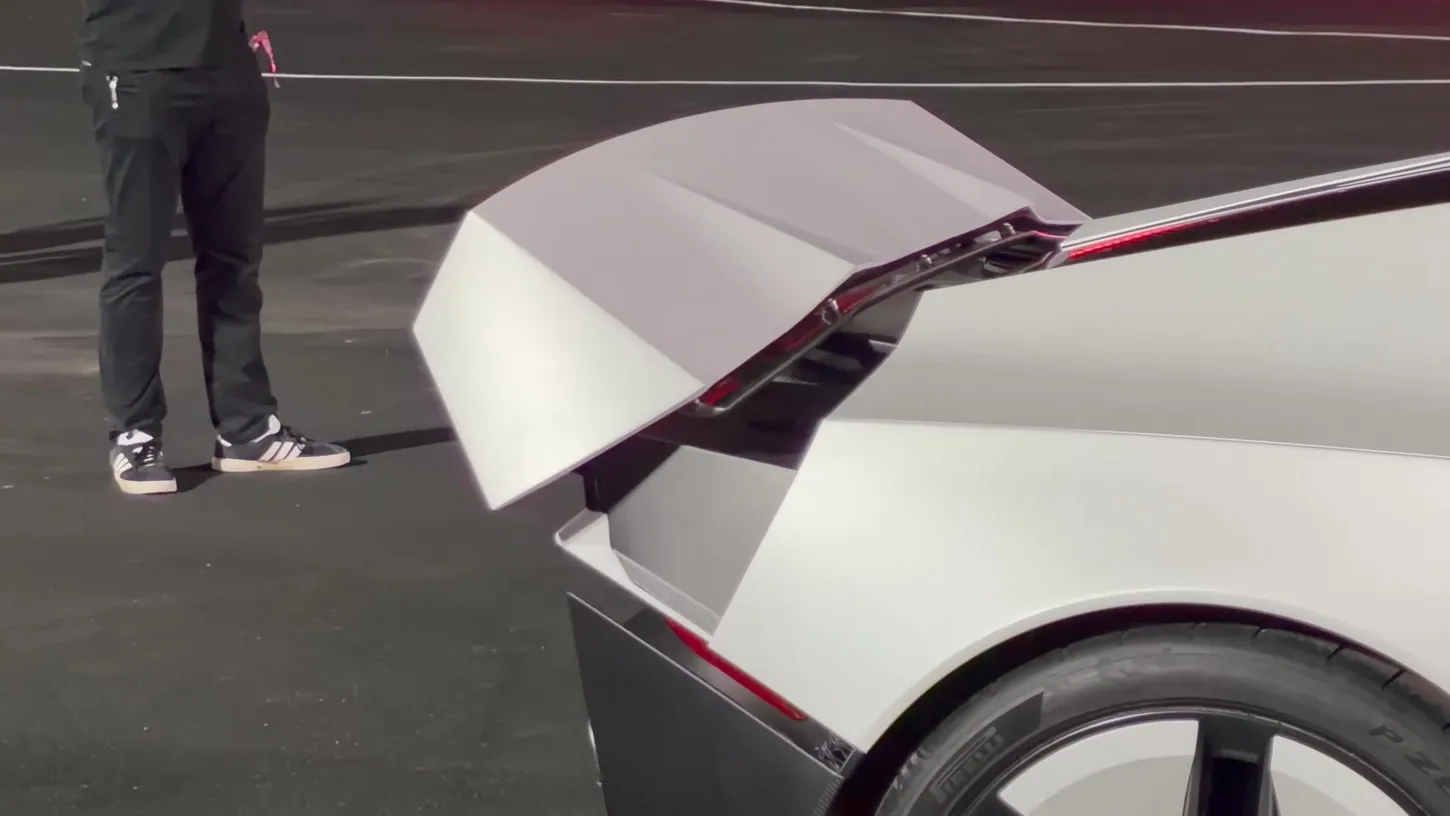 Close-up of a white Tesla Roadster's raised rear wing on its support struts, parked on asphalt at night beside a person in black trousers