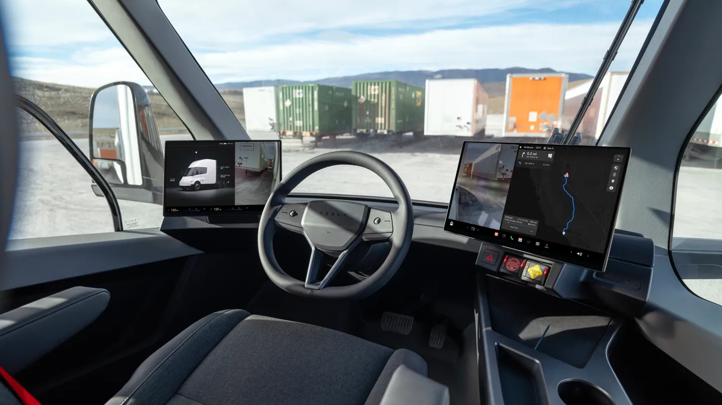 Tesla Semi cab interior seen from behind the center driver's seat, with a touchscreen on each side of the steering wheel and trailers parked outside the windshield