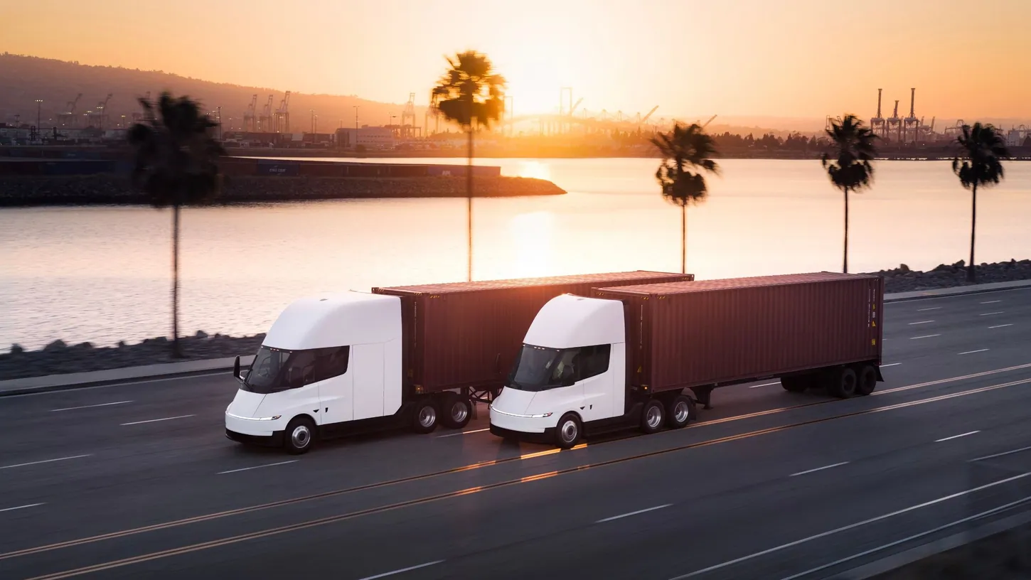 Two white Tesla Semi trucks pulling red shipping containers along a waterfront road at sunset, with palm trees and port cranes behind them