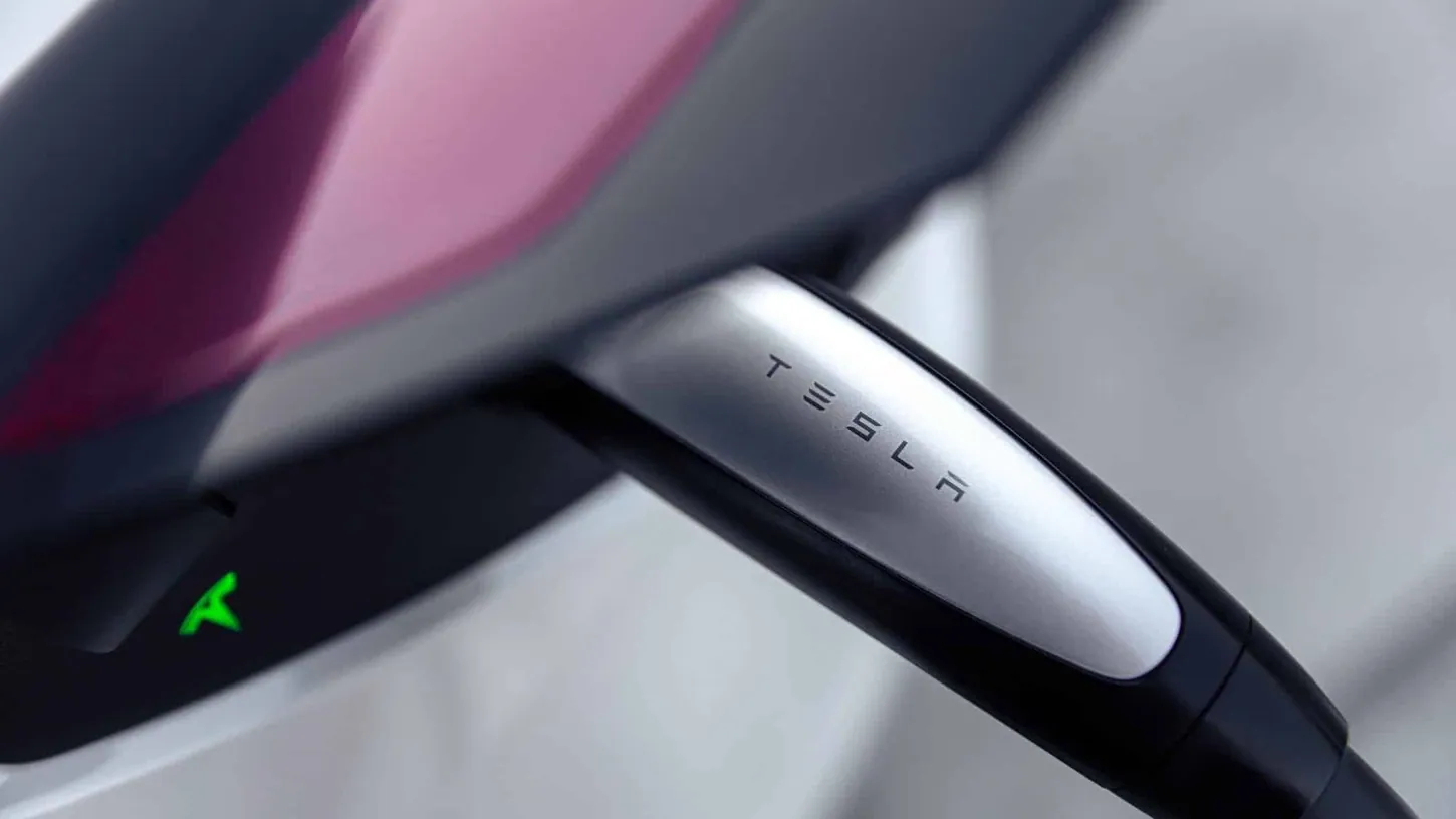 Close-up of a Tesla Supercharger charging handle plugged into a dark red Tesla, with the charge port light glowing green