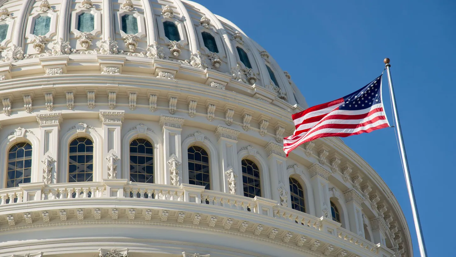 Close-up of the white U.S. Capitol dome with an American flag flying on a pole in front of it against a clear blue sky