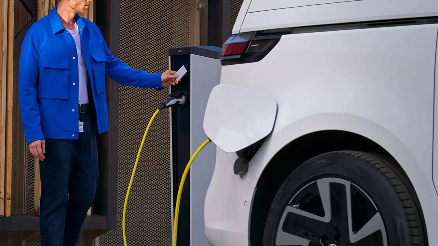 Man in a blue jacket holding a charging card to a charging post connected by a yellow cable to a white Volkswagen ID. Buzz Cargo