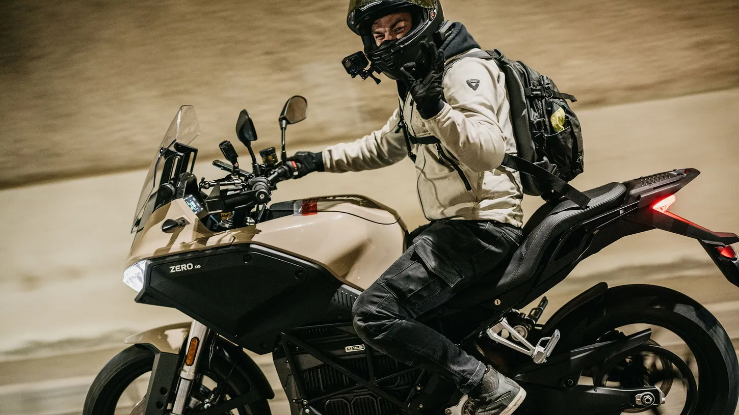 Rider in a light jacket and black helmet waving from a beige and black Zero DS electric motorcycle at speed
