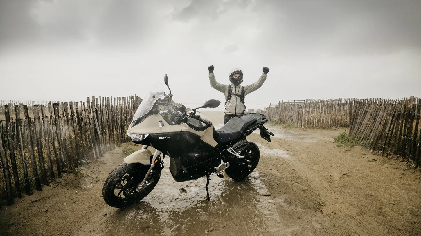Rider with both arms raised behind a beige and black Zero DS electric motorcycle parked in a muddy puddle on a sandy track lined with wooden fencing in heavy rain