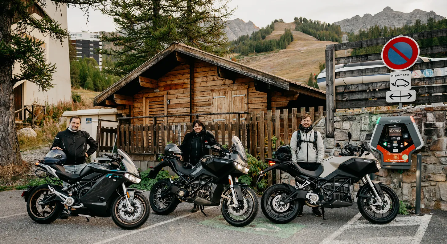 Three Zero electric motorcycles, an SR/S, a DSR/X and a DS, parked beside an EBORN charging station in a mountain village with three riders standing behind them